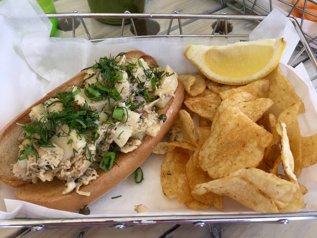 Hearts of palm salad on a sub roll, topped with fresh dill, and a portion of chips with a lemon slice in a metal basket lined with paper.