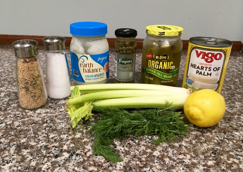 Ingredients for hearts of palm salad are arranged on a marble counter. The ingredients include salt and pepper in shakers, a container of mock mayo, a jar of capers, dill spear pickles, a can of hearts of palm, celery stalks, sprigs of fresh dill, and a lemon.