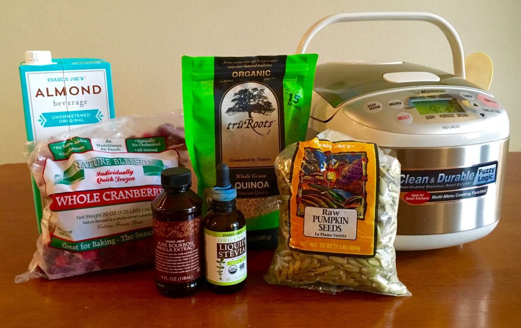 An assortment of ingredients for warm quinoa cranberry breakfast cereal sitting on a wooden table. The ingredients include a rice cooker, raw pumpkin seeds in a bag, a bag of quinoa, a bottle of liquid stevia, a bottle of vanilla extract, a bag of whole cranberries, and a carton of almond beverage.