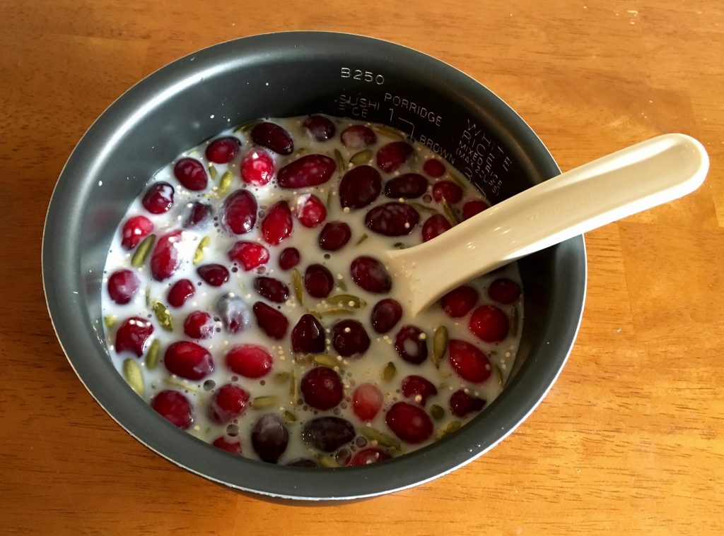 A rice cooker pot with the uncooked ingredients for warm quinoa cranberry breakfast cereal inside of it. There is a white ladle inside the pot and the pot is sitting on a wooden table.