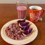 An assortment of foods. On a white plate is slices of a dark pink fruit and warm quinoa cranberry breakfast cereal. There is a red mug with a bird on it containing tea and a glass containing a pink smoothie.