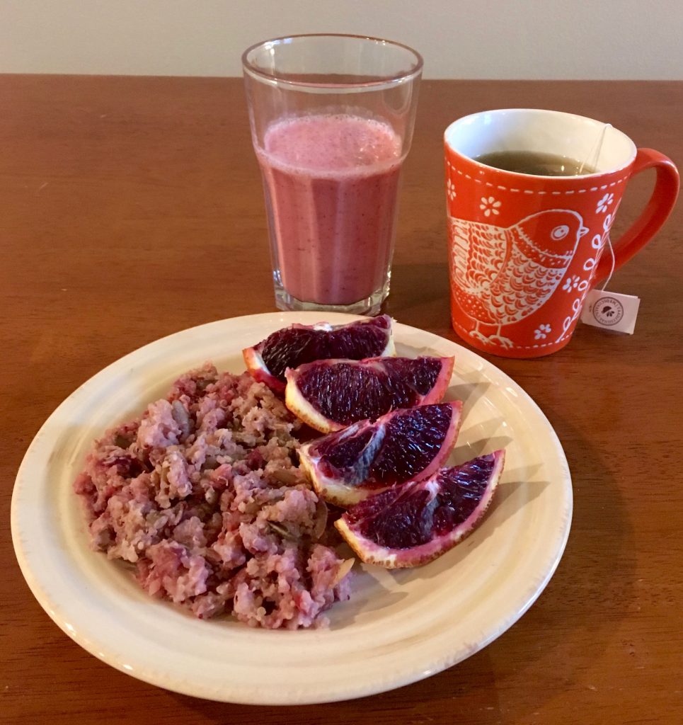 An assortment of foods. On a white plate is slices of a dark pink fruit and warm quinoa cranberry breakfast cereal. There is a red mug with a bird on it containing tea and a glass containing a pink smoothie.
