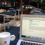 Image from an outdoor café. In the foreground are a vegan tea latte and laptop open to the writing website 750words.com. In the background appear metal chairs and tables inside a metal fence. Beyond the café are cars, trees, and row houses.