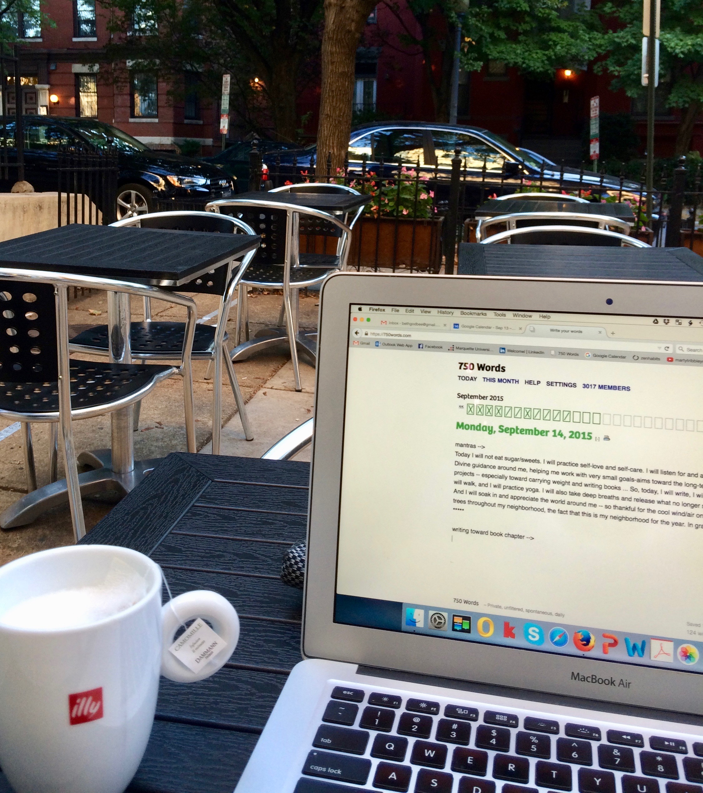 Image from an outdoor café. In the foreground are a vegan tea latte and laptop open to the writing website 750words.com. In the background appear metal chairs and tables inside a metal fence. Beyond the café are cars, trees, and row houses.