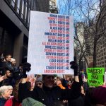 Photo from a crowded protest with a poster in the center reading: "Human Rights are Women's Rights are LGBTQ+ Rights are Native Rights are Black Rights are Latinx Rights are Immigrant Rights are Refugee Rights are Muslim Rights are All Religion Rights are Homeless Rights are Disability Rights are Survivor Rights are Veteran Rights are Elder Rights are Child Rights are Student Rights are American Rights." The poster includes blue and red letters against a white background. Photo credit to Lauren Fitzgerald.