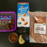 The following ingredients appear against a black table top: (1) Fancy Medjool dates; (2) Almond Breeze unsweetened original almond milk; (3) HealthWorks cacao powder; (4) two avocados, one cut open to see the green inside; and (5) a small metal bowl containing ground cinnamon.