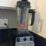 View of kitchen countertops and light-colored cabinets with a black-and-grey Vitamix in the center. Ingredients are inside the blender, ready to be mixed into chocolate avocado pudding.