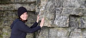 This photo shows Beth in a black fleece jacket and hat, pressing against a stone wall, with head turned to face the camera, and smiling. The rock stretches outward to the right.