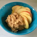 This photo shows a blue bowl against a white tabletop. Inside the bowl is the sweet spread (a light brown color with creamy texture) next to sliced apples (light, yellowish centers with red skin).