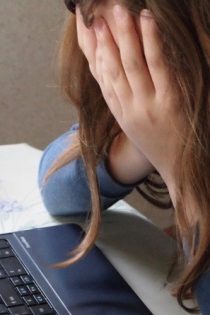 This image shows a person (read as a white woman) with hands and hair covering the face and whole body slumped in front of an open laptop.