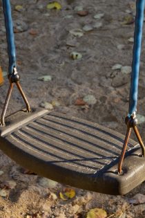 This image shows a brown swing seat held by two blue rubber cords. Sandy dirt with fallen leaves are in the background.