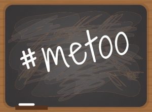 This images shows the #metoo hashtag in white script printed diagonally on a blackboard with messy erasure marks in the background.