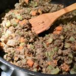 This photo shows the cooking in-action: a mix of lentils, carrots, celery, and onions are in a frying pan, being stirred by a wooden spatula.