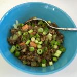 This photo shows the final meal: a mixture of lentils and veggies topped with green onions—in a blue bowl against a white tabletop.