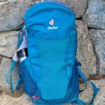 Here a teal-blue hiking backpack (lighter day-pack with thermos visible) sits against a stone bench.