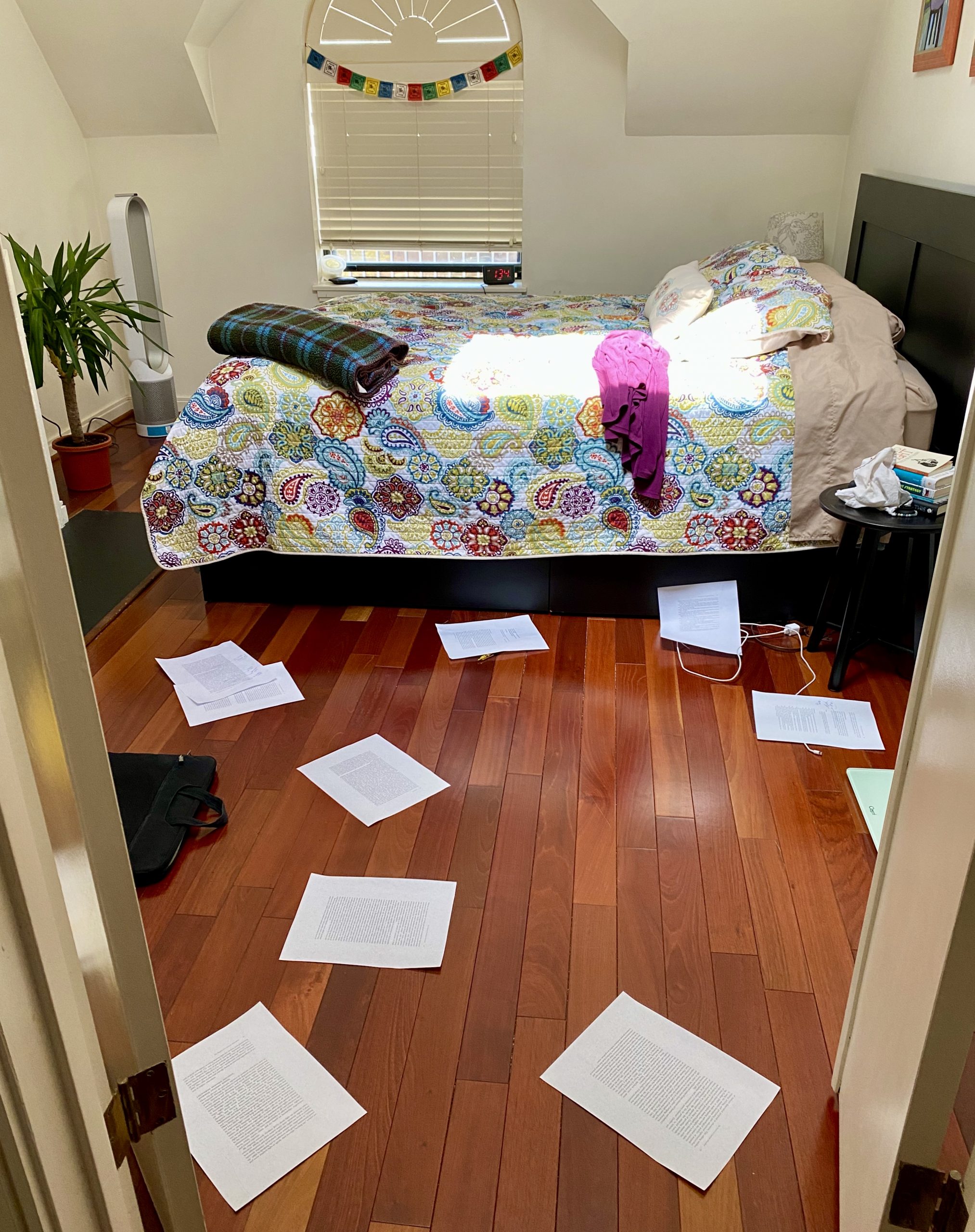 This photo shows scattered papers (page proofs) across a wooden floor in a bedroom—with a colorful bedspread, pillows, blanket, sweater, plant, air purifier, bedside table, books, window, and white walls all part of the scene.