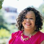 This photo shows Tamika Carey, Ph.D. smiling, wearing a pink blouse, and standing outdoors (surrounded by green leaves and a building blurred in the distance).