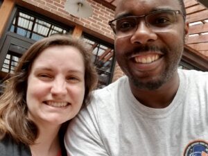 This selfie shows Beth Godbee on the left and Cedric Burrows on the right: both smiling—positioned in front of a brick building and glass windows reflecting steel beams.