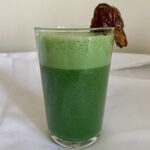 This image shows the spirulina shake (a bright green smoothie) in a clear glass, a brown date attached to the rim, against a white tablecloth.