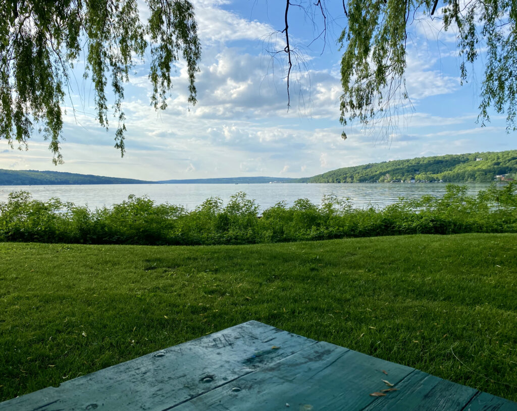 This photo shows a late summer afternoon. The corner of a green picnic table points toward a green field and blue lake stretching out to blue-green hills. Leaves hang down, and puffy white-grey clouds float overhead. The scene, hopefully, captures the possibilities of summer: open, outdoor space inviting contemplative practices.