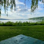 This photo shows a late summer afternoon. The corner of a green picnic table points toward a green field and blue lake stretching out to blue-green hills. Leaves hang down, and puffy white-grey clouds float overhead. The scene, hopefully, captures the possibilities of summer: open, outdoor space inviting contemplative practices.