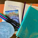 This photo shows multiple journals, open to a lined notebook page that says “contemplative writing.” A colorful divination card, blue crystal, and three markers sit on top of the journals.
