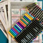 This image shows my current journaling and drawing supplies: colorful markers and pastels on top of loose papers with handwriting and a 12-month project vision.