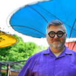 A photo of David smiling looking forward toward the camera. He's wearing a purple guayabera shirt with chunky Black plastic circle frame glasses. David has a silver nose hoop ring. His hair and beard are a mix of dark and light gray. The background consists of green foliage next to and behind David with three circle canopy structures overhead. One canopy is yellow, another blue, and the third red.