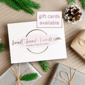 This image shows a scene of wrapped packages, a pine cone, and evergreen branches. A white text box shares the circular logo for Heart-Head-Hands: Everyday Living for Justice, and another text box reads: “gift cards available.”