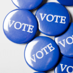 This photo shows several blue buttons with VOTE printed in white capital letters. The buttons are overlapping and against a white background.
