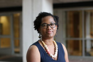 This professional headshot shows Candace Epps-Robertson, Ph.D. with glasses, earrings, beaded necklace, and navy wrap dress. Candace is standing outside a building in contrasting light and shadow.