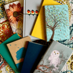Pictured here are eight colorful journals -- with designs that include trees, flowers, and birds -- spread across a patterned bedspread.