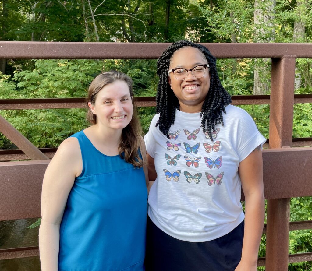 This photo shows Beth and Candace on a walk together—standing side-by-side, smiling, and wearing summer shirts (blue tank and butterfly t-shirt). We’re standing on a bridge surrounded by nature (green trees and leaves).