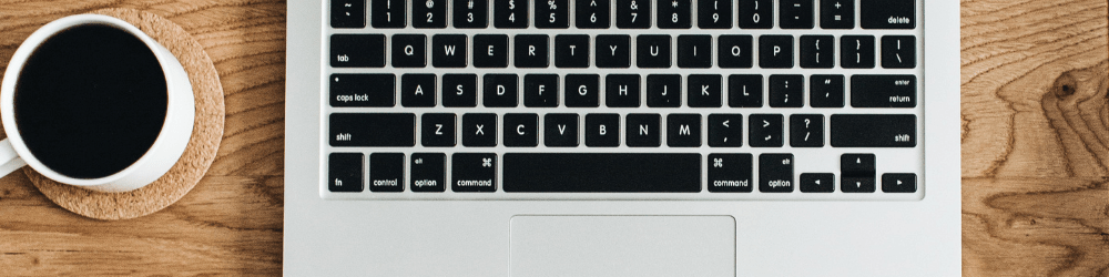 A large rectangular image showing a black computer keyboard and a cup of black coffee in a white mug, sitting on a coaster. The computer and coffee cup are on a wooden table.