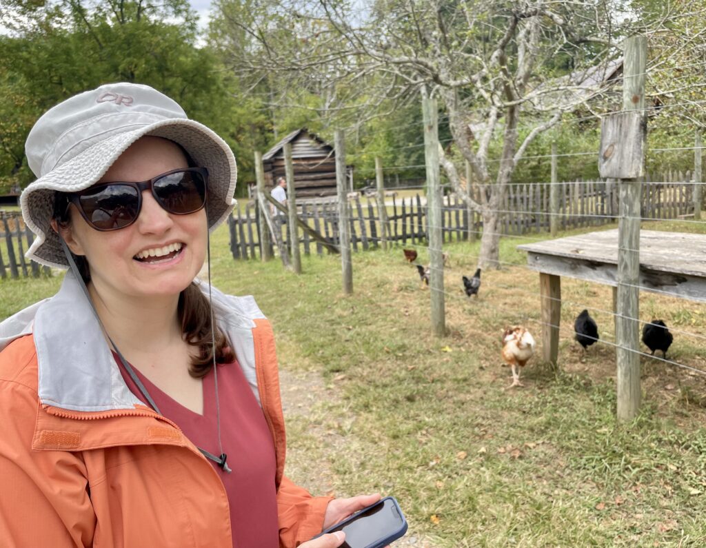 Beth stands in front of a fenced in patch of farm land with chickens. She is wearing an orange rain jacket, a red shirt, brown sunglasses, and a beige hat.