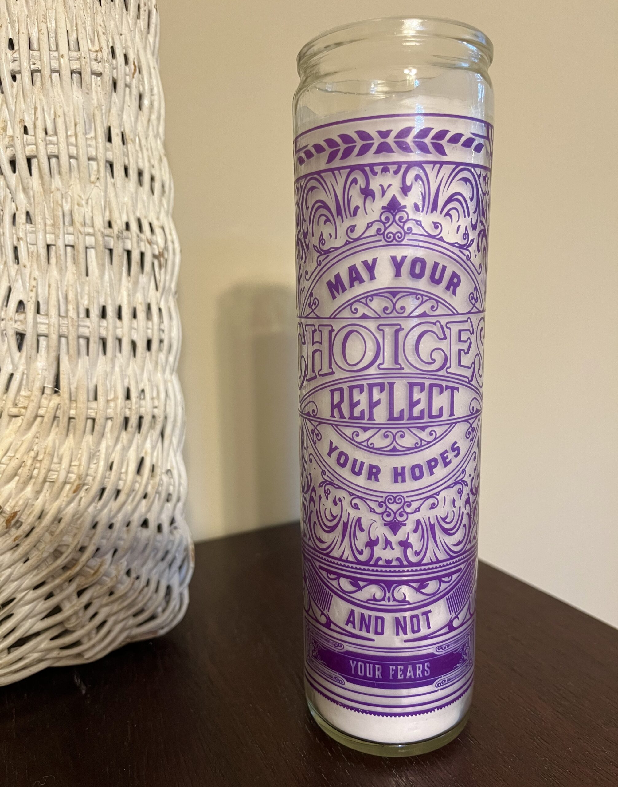 A tall, white candle stands in a glass container with purple detailing on the front. The candle's cover reads, "May your choice reflect your hopes and not your fears." The candle sits on a wooden surface in front of a white wall.
