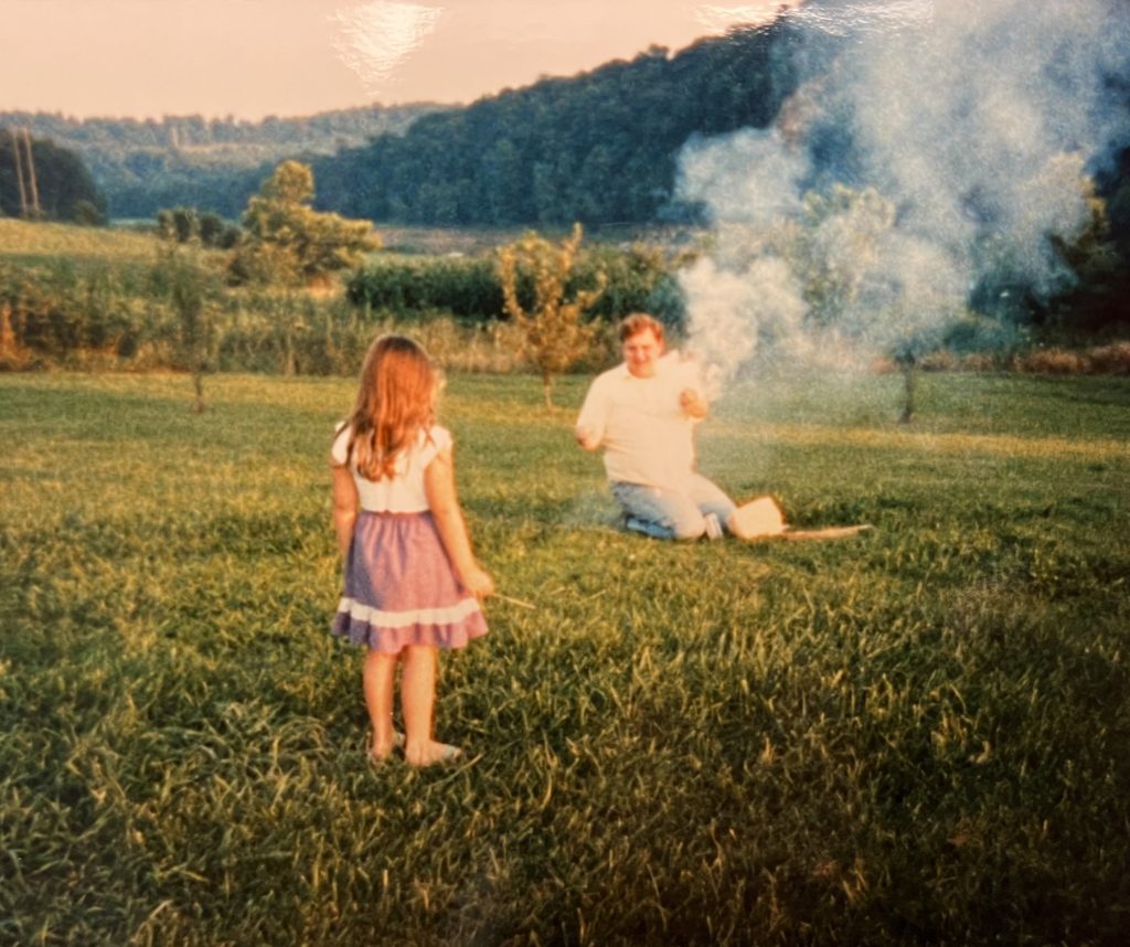 Beth in her childhood years stands in a purple skirt and white shirt in a grassy, mountain surrounded field near her father who holds smoking fireworks.
