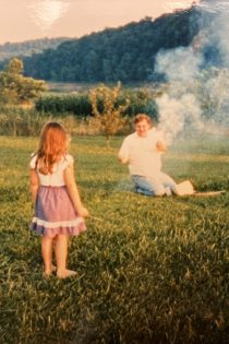 Beth in her childhood years stands in a purple skirt and white shirt in a grassy, mountain-surrounded field near her father who holds smoking fireworks.