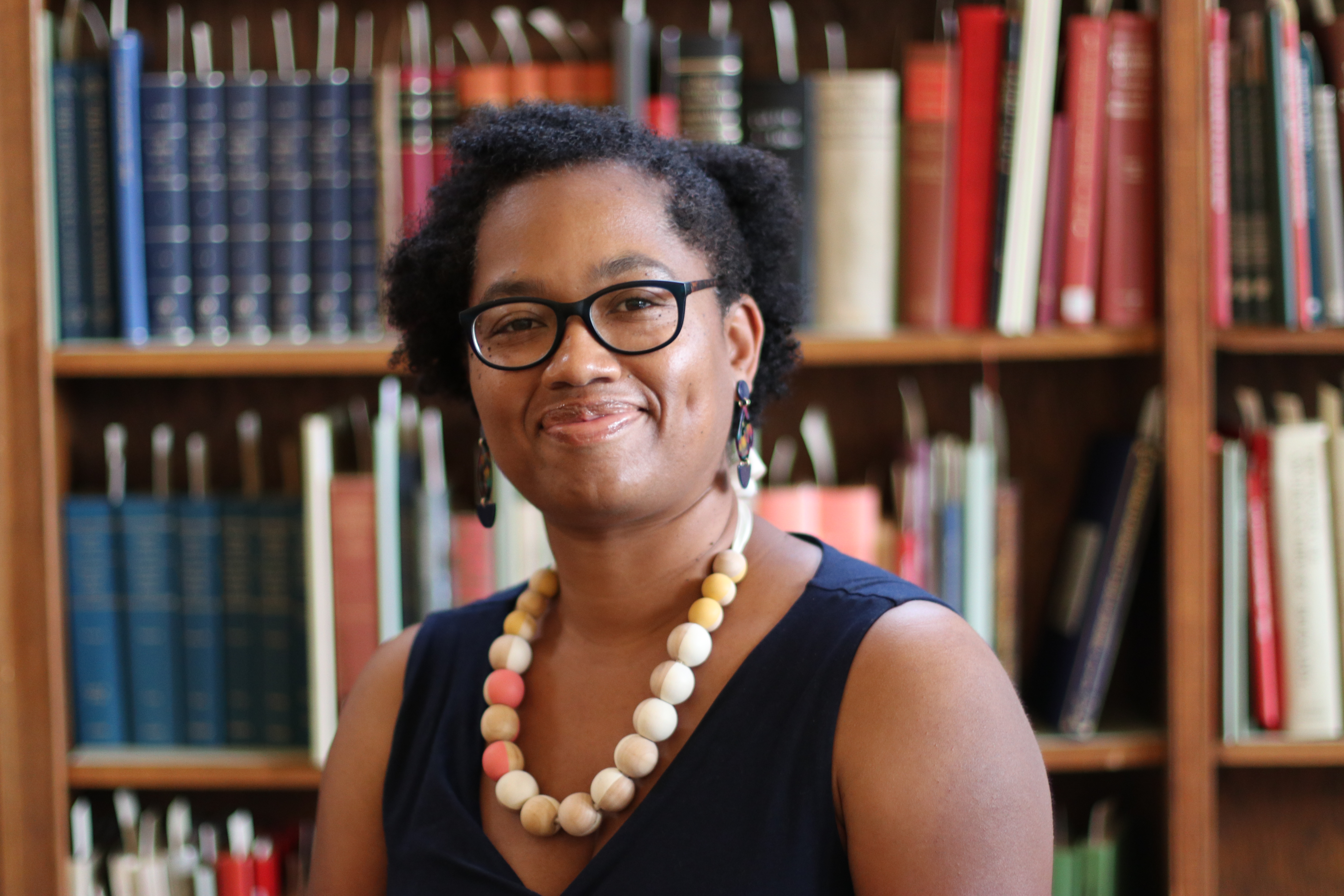 Dr. Candace Epps-Robertson sits smiling in front of a bookshelf covered in books, dressed in a navy blue sleeveless dress and a large, beaded necklace. She wears black glasses and also multi-colored earrings.