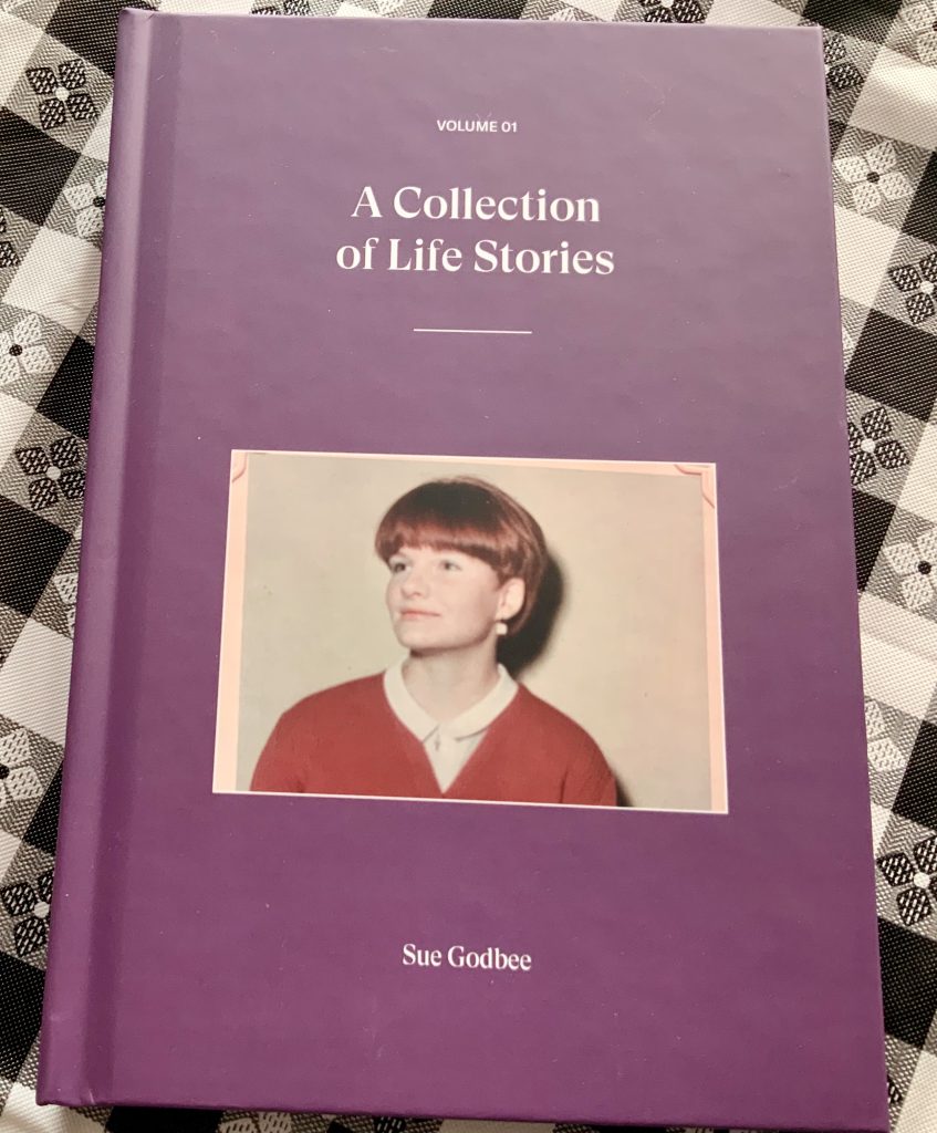 A purple book with a picture of Beth's mother on the cover lays on a black and white checkered tablecloth. The book is titled "A Collection of Life Stories" and the author's name, Sue Godbee, is written below the image that takes up the lower space on the book's cover. This is volume 01.