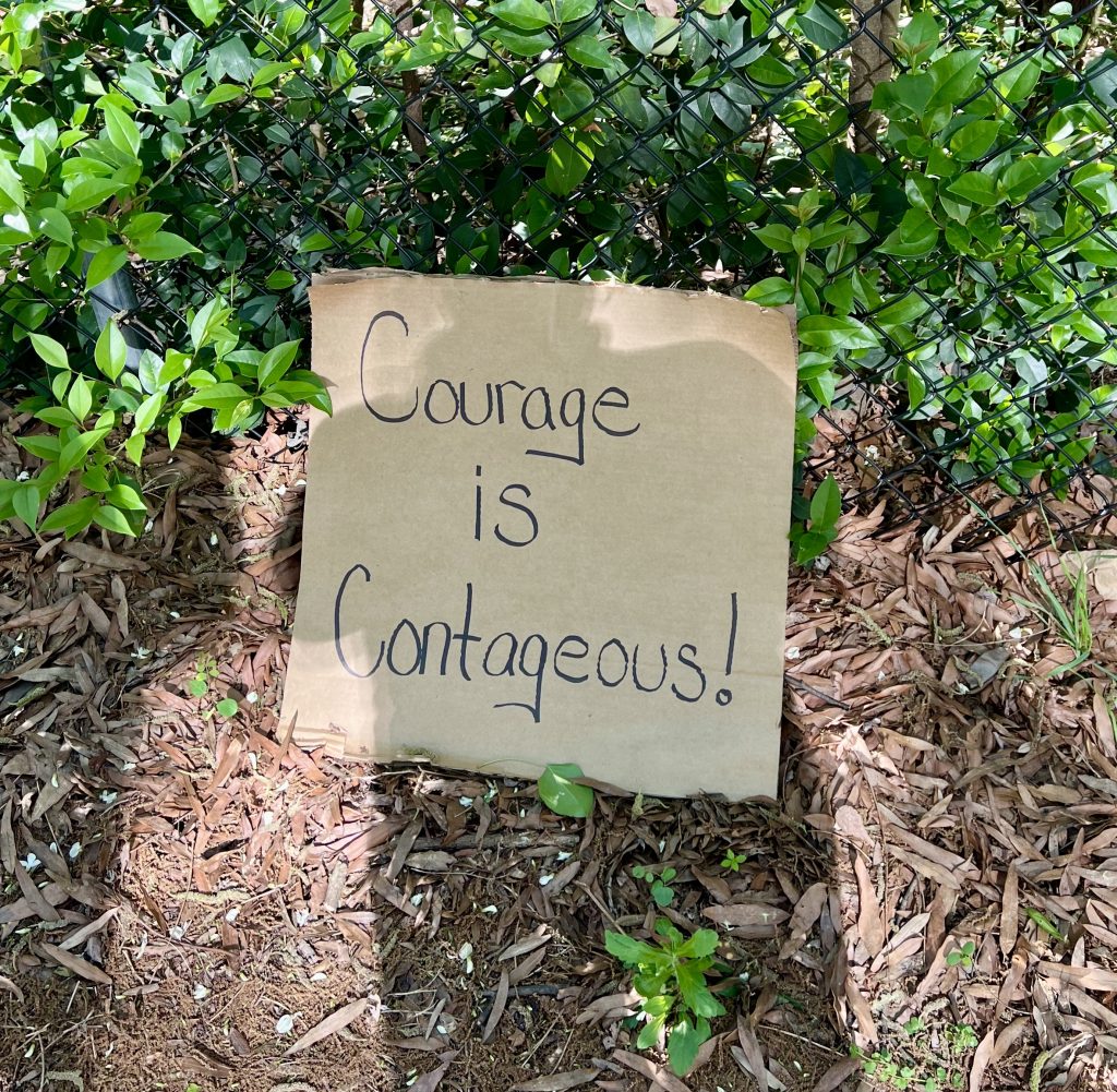 A cardboard sign leaning against a foliage covered fence and a mulch covered ground reads "Courage is Contageous!" Candace Epps-Robertson, Ph.D. of Full Circle Writing, LLC. Candace took this photo of a handwritten sign that asserts: "Courage is contagious!" Check out Candace's offerings here: https://full-circle-writing.com/
