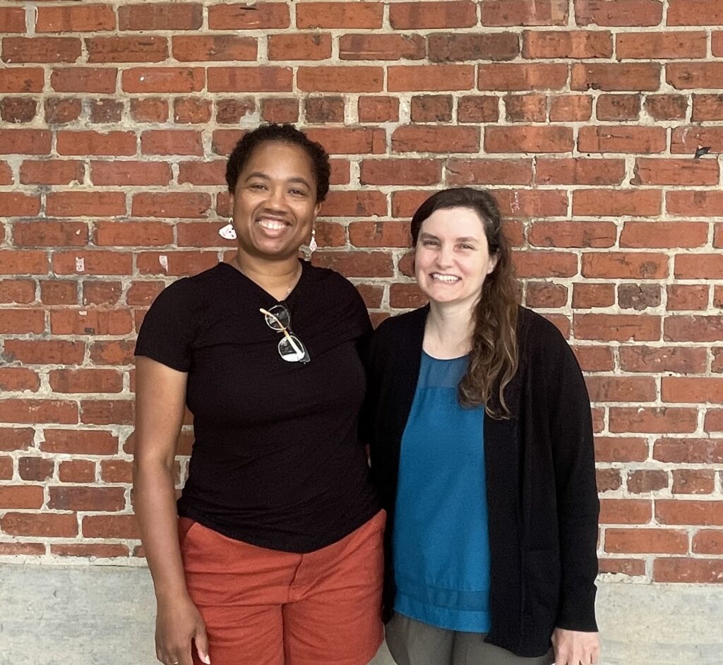 A photo of Beth and Candace standing next to each other in front of a brick wall. Candace is wearing a black shirt, white earrings, and orange shorts. Beth is wearing a black sweater, blue shirt, and green pants. Both smile at the camera.