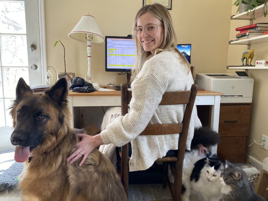 Sarah Gettel sits at her desk wearing a white sweater and clear-rimmed glasses. Turning back to look at the camera, she smiles while petting her large dog, Wilbur, while her two cats, Puck, a black and white cat; and Atticus, a grey cat; sit next to her chair.