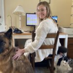 Sarah Gettel sits at her desk wearing a white sweater and clear-rimmed glasses. Turning back to look at the camera, she smiles while petting her large dog, Wilbur, while her two cats, Puck, a black and white cat; and Atticus, a grey cat; sit next to her chair.