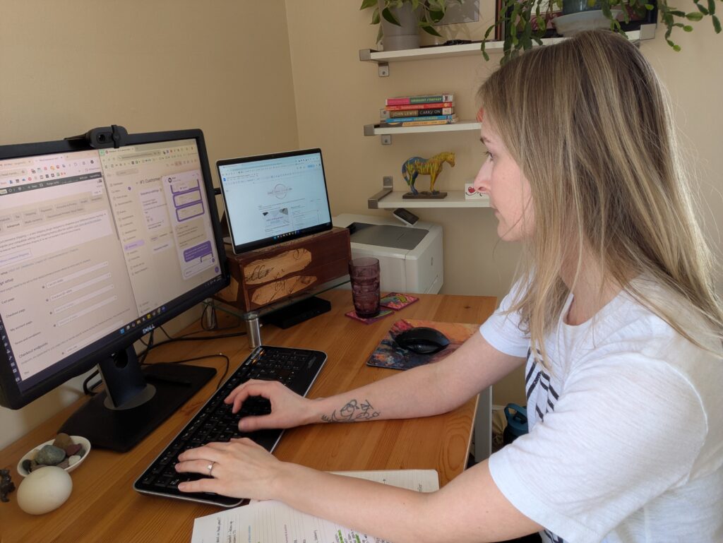 Sarah Gettel, wearing a light grey t-shirt, works at her desk, the surface of which is a warm-brown tone of wood. She has two monitors open. One monitor shows a split screen of different websites. The second monitor shows the Heart-Head-Hands website.