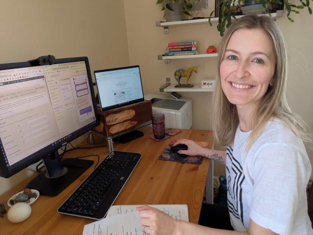 Sarah Gettel, wearing a light grey t-shirt, smiles at the camera from her desk. On top of her wooden desk is a computer setup with two monitors, a keyboard, and an assortment of small crystals in a dish. Behind her, white shelves contain books, plants, and a small horse figurine.