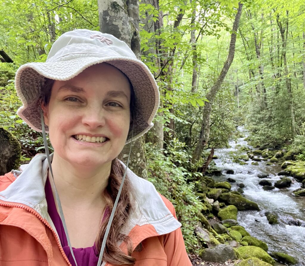 Beth, wearing a green hat, maroon shirt, and orange jacket, smiles at the camera next to a flowing stream of water and green trees in the Great Smokey Mountains.