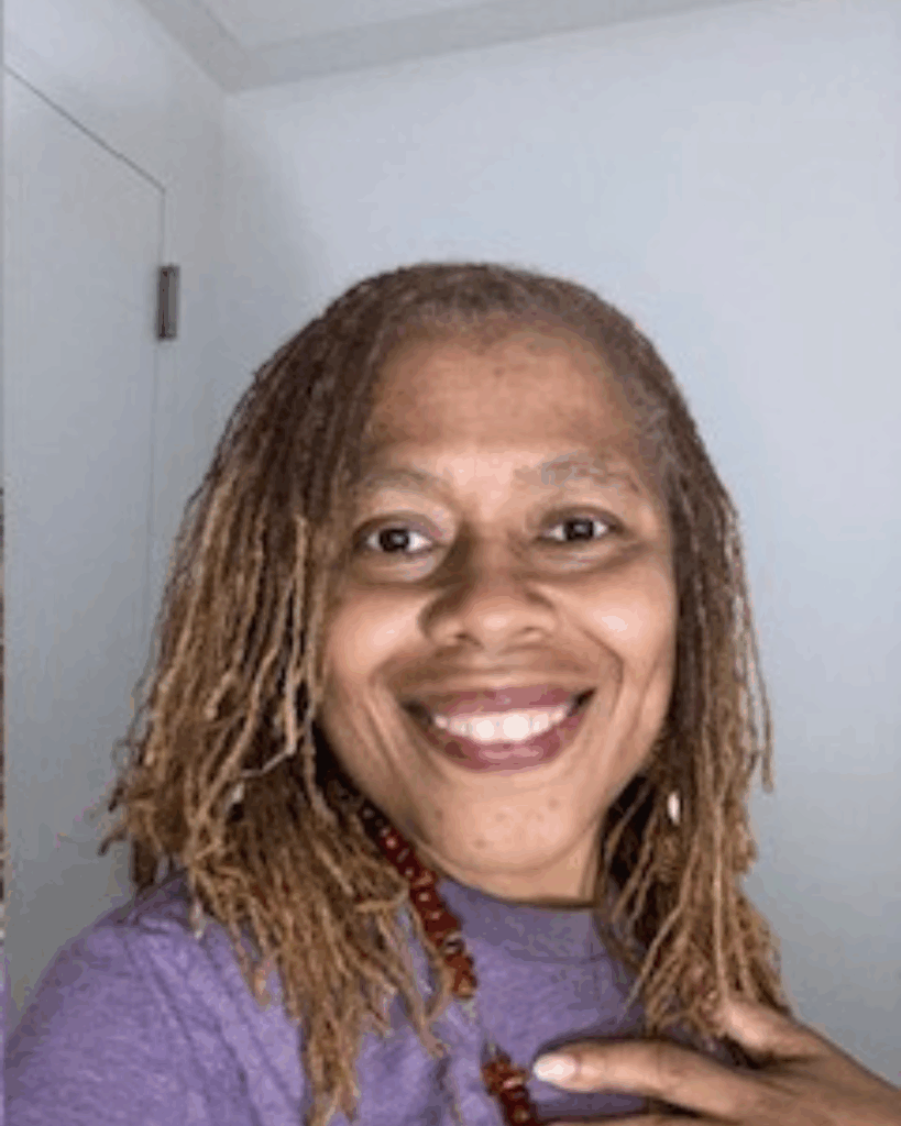 A photo of Pam Jones smiling at the camera in a purple sweater and red stone necklace.