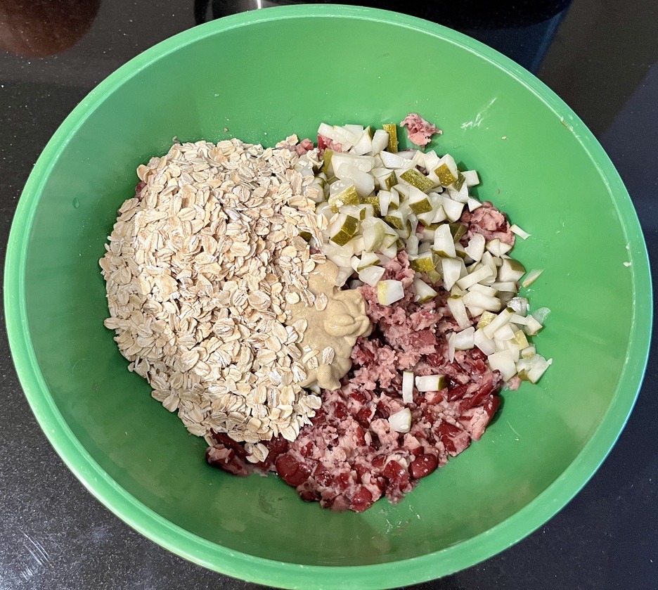 A green bowl contains the prepared ingredients needed to make veggie burgers. In the bowl rolled oats, dijon mustard, chopped pickles, and mashed red beans sit together unmixed.