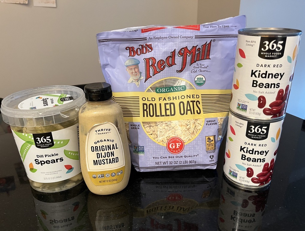 A small stack of ingredients to make veggie burgers sit on a black table surface. The ingredients include 2 cans of dark red kidney beans, old fashioned rolled oats, dijon mustard, and dill pickle spears. The ingredients sit on the table, closely pushed together in the order they were listed in from right to left.